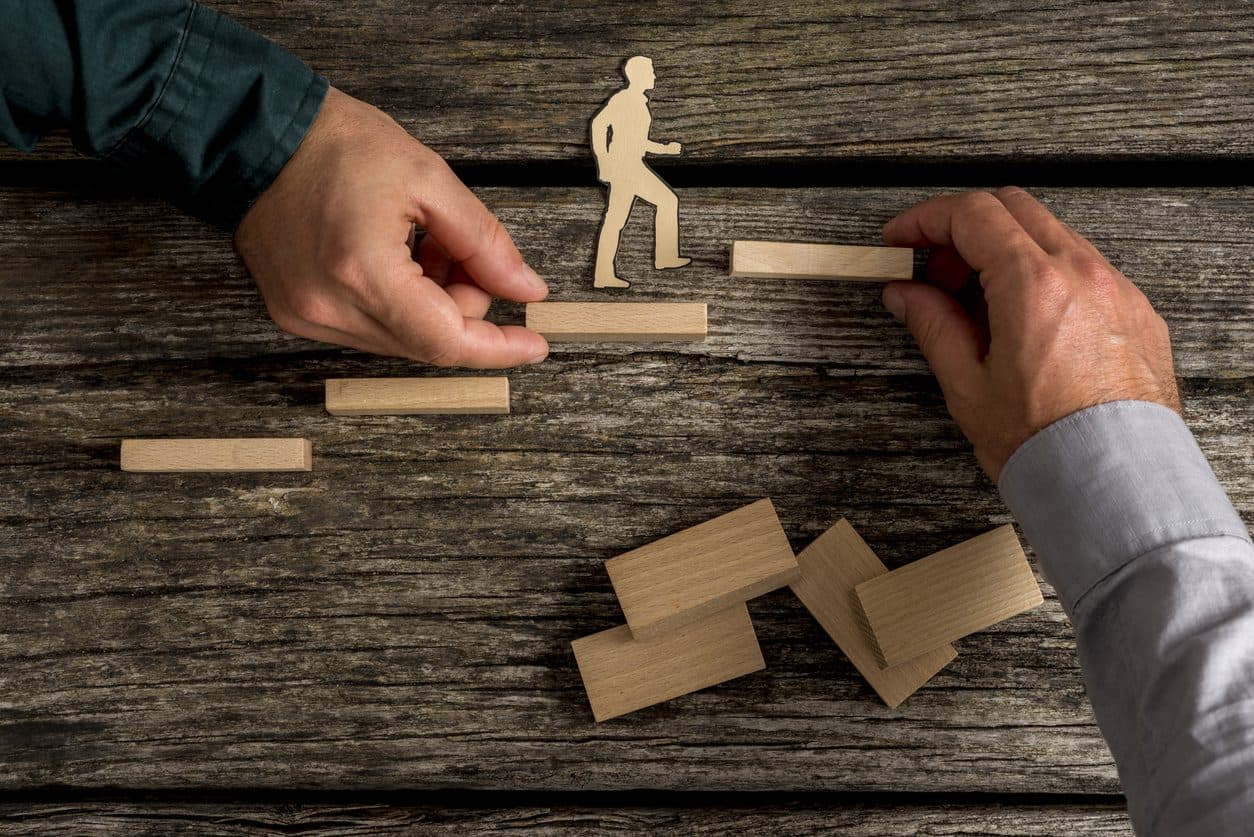 Two unidentifiable adult people building small toy wood staircase as small wooden person walks up the staircase.