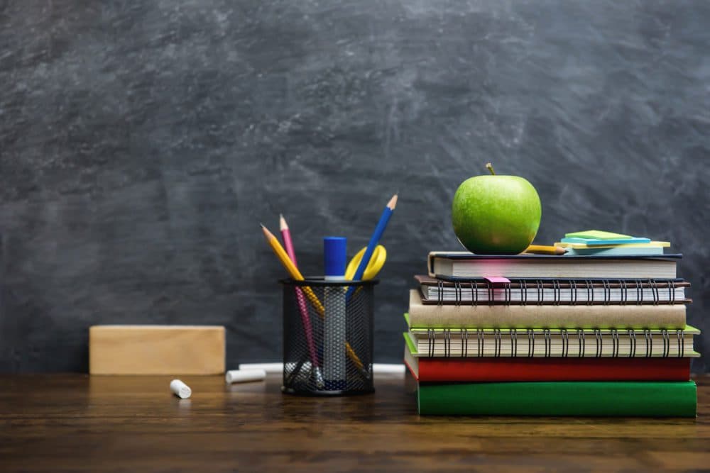 Books, stationery and education supplies on wooden desk in classroom with blackboard in background